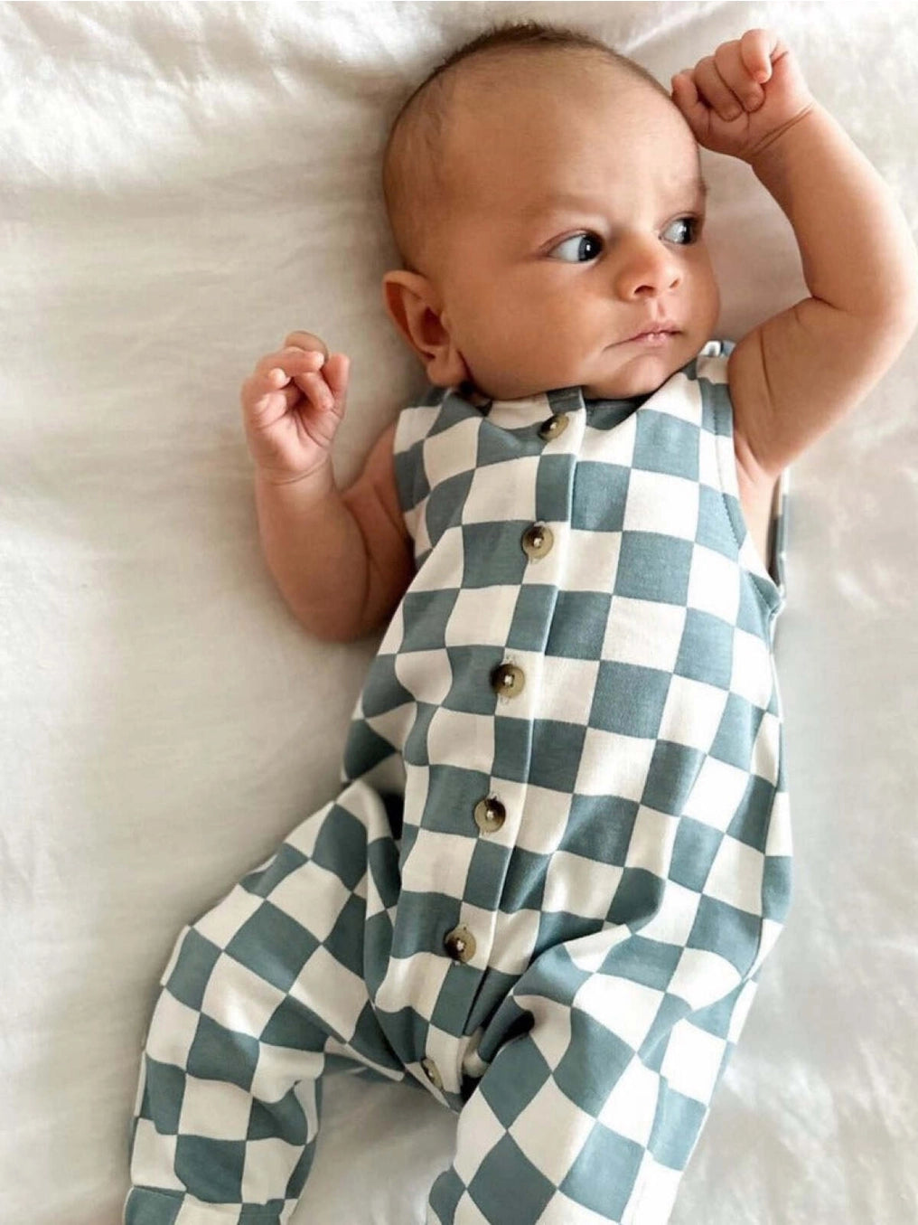 A baby lying on a white surface wearing a blue and white checkered jumpsuit with buttons down the front.