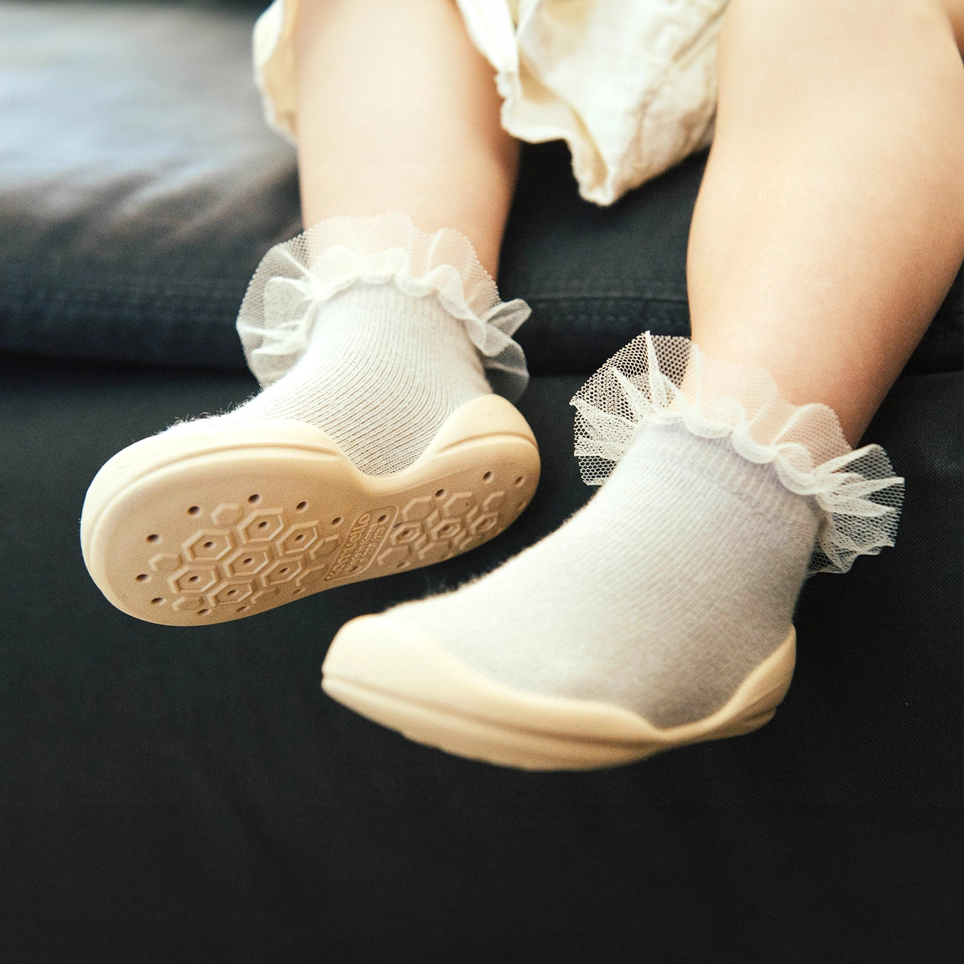 A pair of Komuello toddler shoes with tulle trim on the top, in a beige color, with non-slip soles.