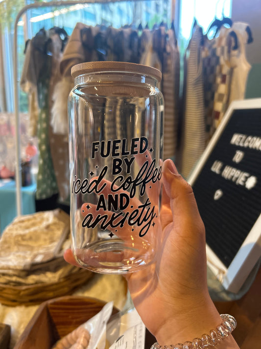 A hand holding a clear glass coffee cup with a bamboo lid, and the text 'Fueled by ice, coffee, and anxiety' printed on it.