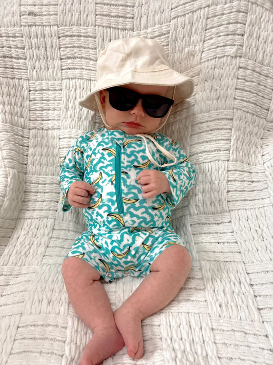 Baby wearing a turquoise swimsuit with a wave pattern and sunglasses, lying on a textured surface.