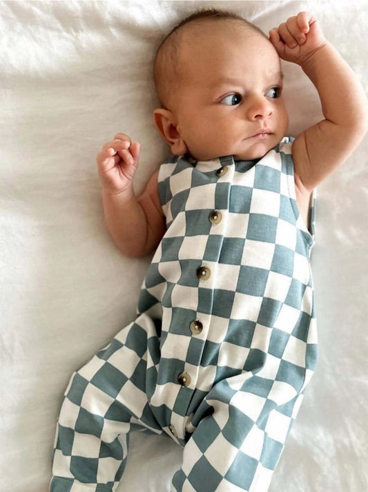 A baby lying on a white surface wearing a blue and white checkered jumpsuit with buttons down the front.