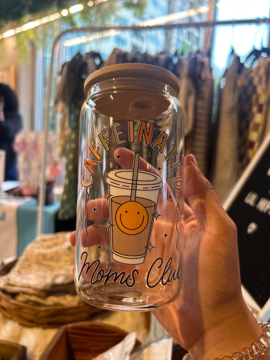 A hand holding a 16 ounce glass coffee cup with a bamboo lid and a graphic that says 'Caffeinated Moms Club' along with a smiley face.