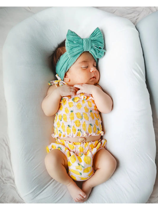 Newborn baby in a yellow and white outfit with a green headband, lying on a white pillow.