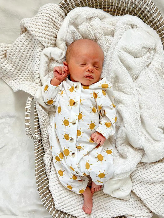 A sleeping newborn baby wearing a long sleeve jumpsuit with yellow sun patterns, lying on a white knit blanket.