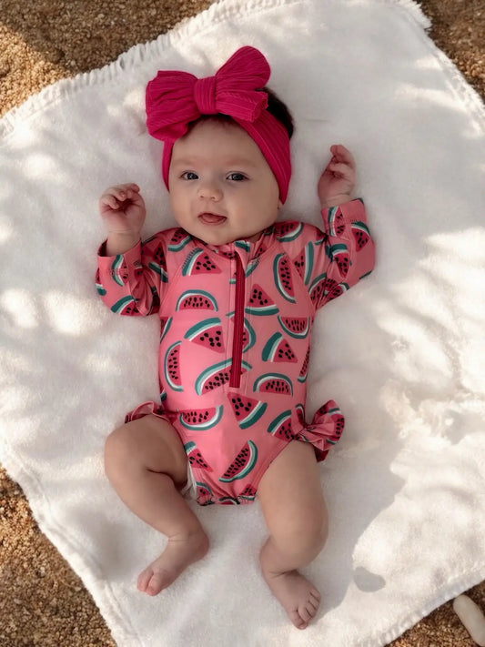 Baby wearing a pink watermelon-patterned onesie and red headband on a white blanket.
